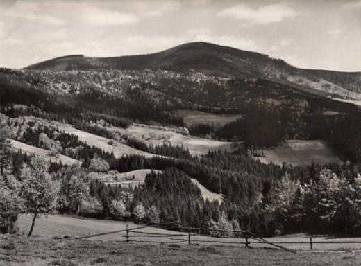 Pohled ze svahu Ostré hory na Lysou horu přes pečlivě obdělané plužiny pasek U Zbuja, Za Ostrou a dalších. Rudolf Janda pořídil tuto fotografii 9. května 1946 a zachytil tak jarní efekt kvetoucích ovocných stromů kolem pasek i svěží zeleně listnatých stromů v pásu smíšeného lesa pod Lysou. 